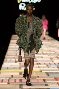 Model walks the runway in a green plaid oversized jacket over striped shorts, with black sandals and a small brown handbag.