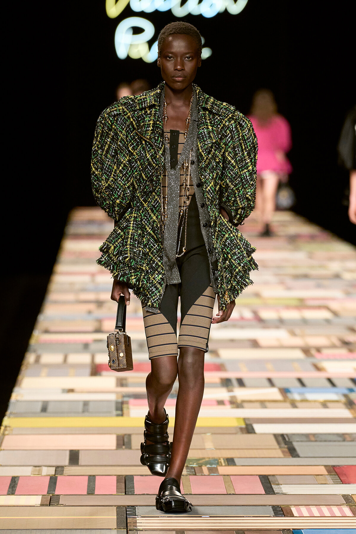 Model walks the runway in a green plaid oversized jacket over striped shorts, with black sandals and a small brown handbag.
