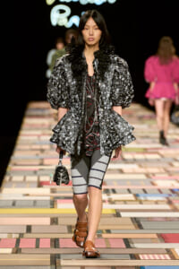 Model walking a colorful tiled runway in a black floral oversized blouse with voluminous sleeves, layered with a dark top and gray striped leggings, carrying a small black quilted bag and wearing brown sandals.