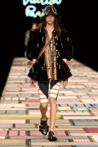 Model struts the runway in a black velvet jacket with silver pins, layered necklaces, a striped top and color-block shorts, and a black embellished hat and sandals.