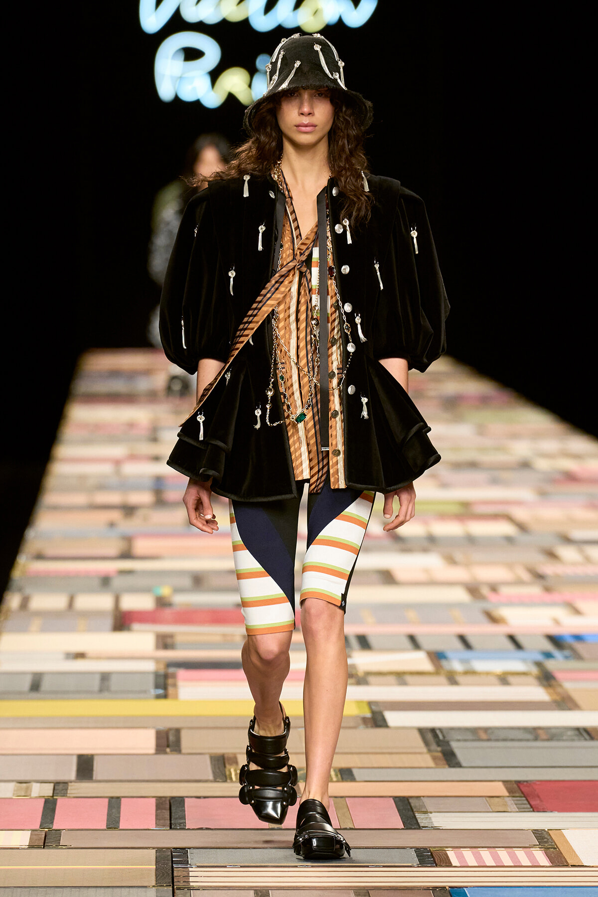 Model struts the runway in a black velvet jacket with silver pins, layered necklaces, a striped top and color-block shorts, and a black embellished hat and sandals.