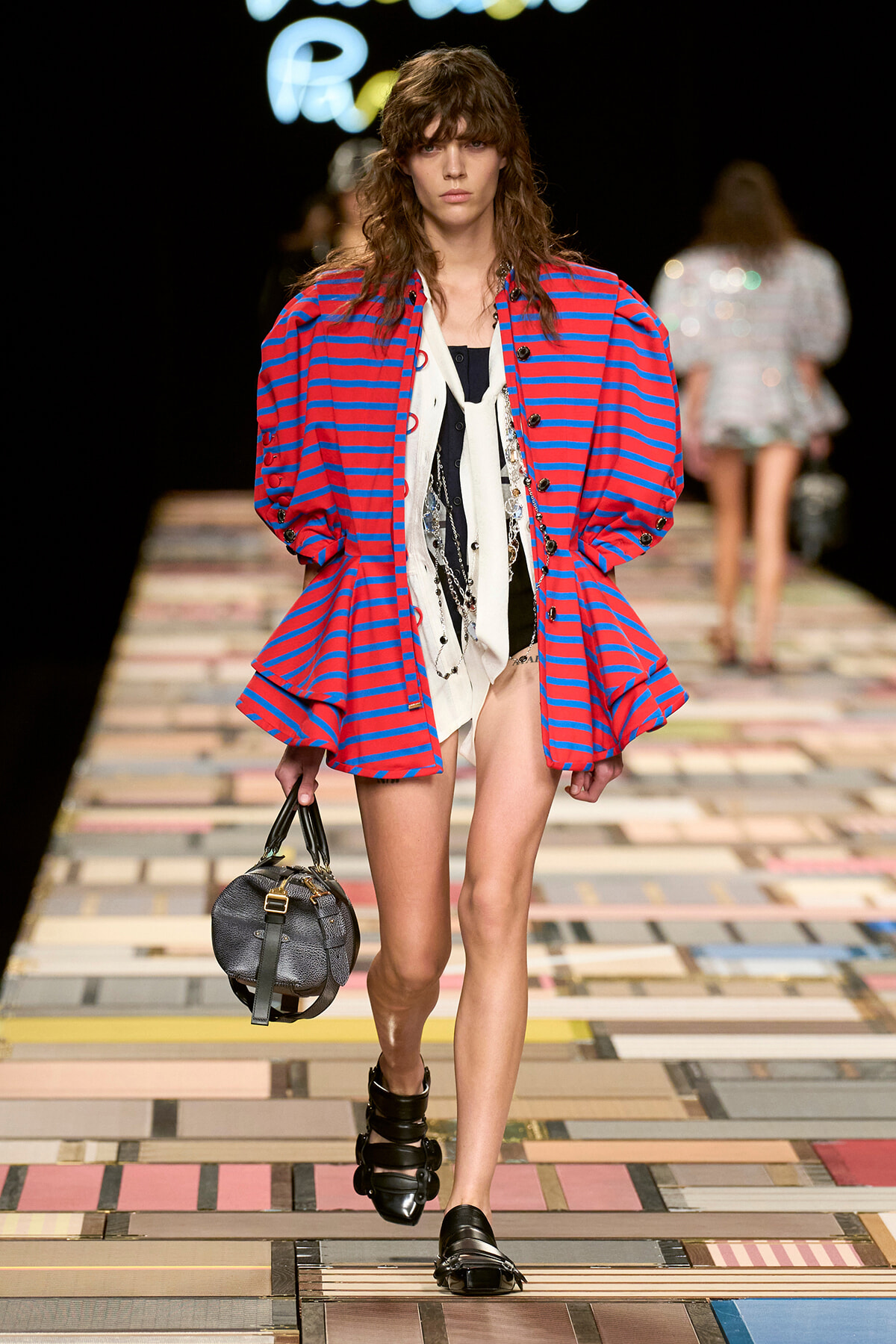 Model on a fashion runway wearing a red-and-blue striped oversized jacket, layered top, and black sandals, carrying a round black bag.
