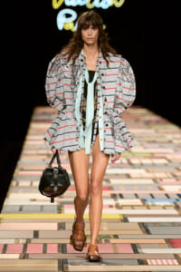 Model walking a fashion runway in a gray striped oversized jacket with puff sleeves, mint scarf, black top, brown sandals, and a black rounded handbag.