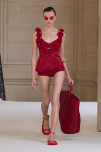 Model on a runway wearing a red rose-embellished romper with matching shorts, carrying a large red tote and wearing red sandals and sunglasses.