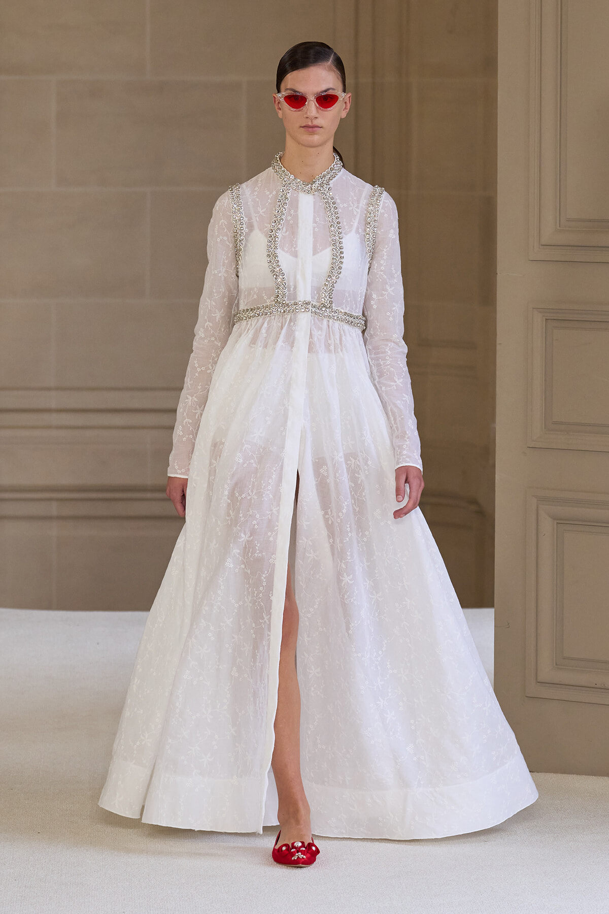 Model in a white lace gown with beaded trim and sheer sleeves on a runway, paired with red sunglasses and red flats.