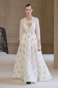 Model on a runway wearing a sheer white gown with gold leaf embroidery and a heart-shaped design on the bodice, long sleeves, and a tied waist ribbon.