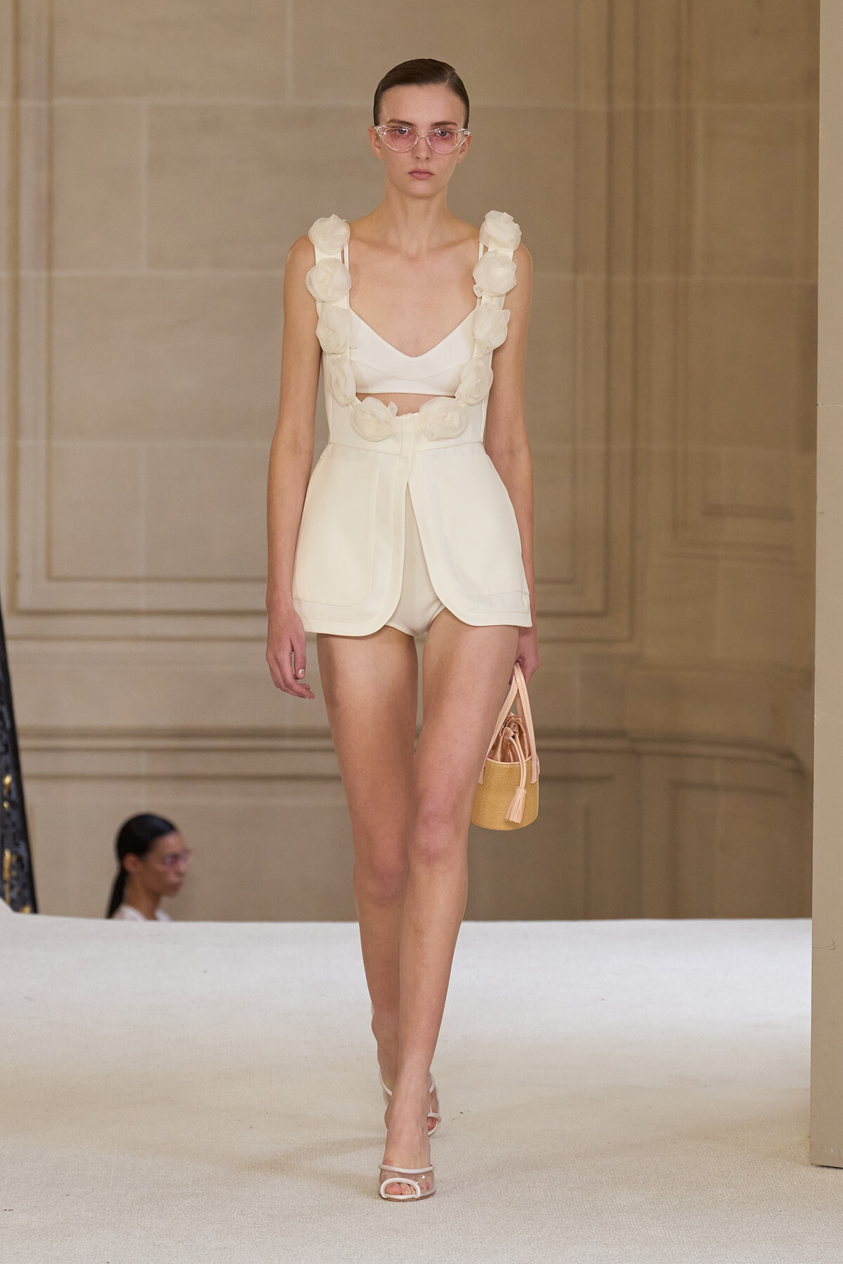 Model walks a runways wearing an ivory two-piece with large fabric flower rosettes on the straps and matching high-waisted shorts; carrying a small straw bag.