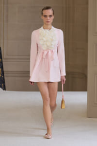 Model on runway in pale pink mini dress with white flower appliqués at the chest, tied waist, and pink flats, carrying a small gold handbag.