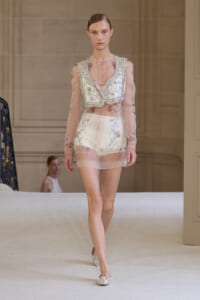 Model walking a runway in a sheer pink floral mini dress with a beaded, silver-embellished vest top and matching pale pink flats.