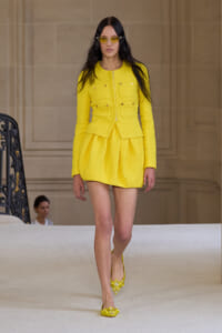 Model walking a runway in a bright yellow tailored blazer with a matching pleated mini skirt and yellow heels with pearl accents. She wears yellow sunglasses and has long dark hair.