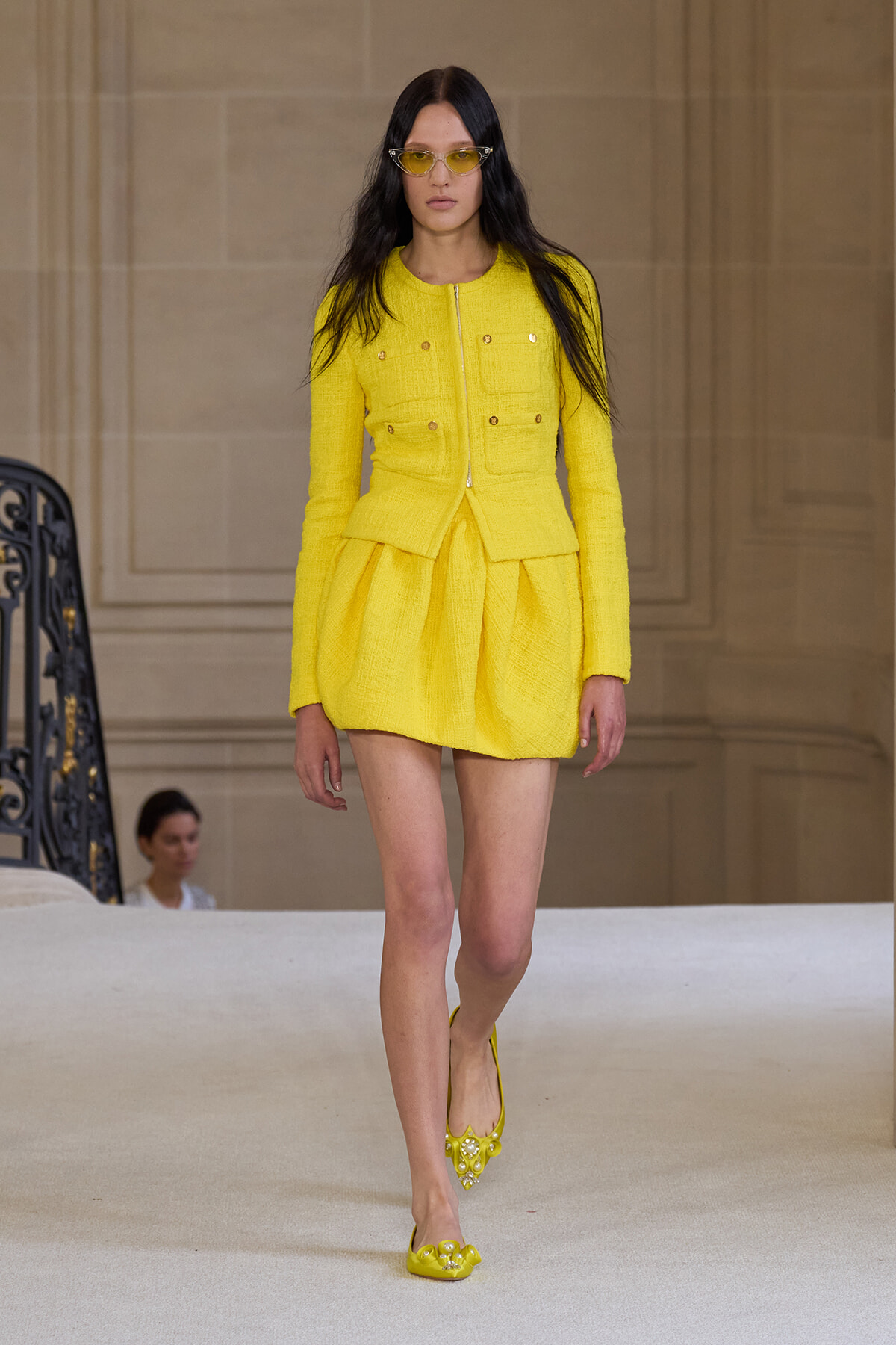 Model walking a runway in a bright yellow tailored blazer with a matching pleated mini skirt and yellow heels with pearl accents. She wears yellow sunglasses and has long dark hair.