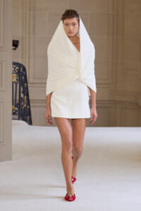 Model on a runway wearing a white draped wrap over a mini dress with red heels.
