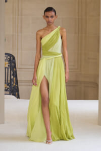 Model walking a runway in a lime-green one-shoulder draped gown with a high slit and white sandals.