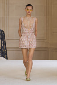 Model walking runway in a pink patterned sleeveless mini dress with sheer lace top and yellow sunglasses, green flats visible