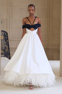 Model walks a runway in a black off-shoulder bodice with criss-cross straps and a white A-line gown with a feathered hem.