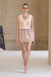 Model walking a runway in a pale pink knit sleeveless top with gold embroidery and a pink patterned mini skirt, wearing orange-tinted sunglasses.