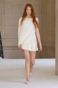 Model walking a runway in a white draped one-shoulder mini dress with soft pleats and metallic flats.