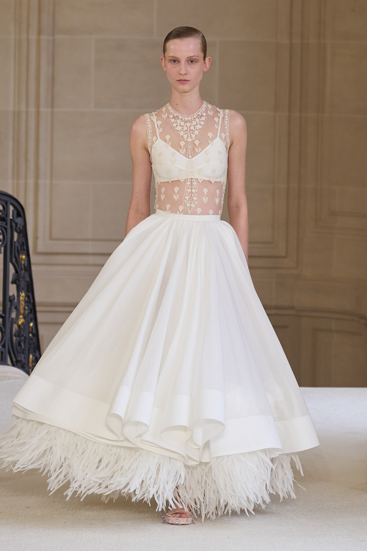 Model wearing a white wedding gown with sheer lace bodice and full skirt, feathered hem visible at the bottom.