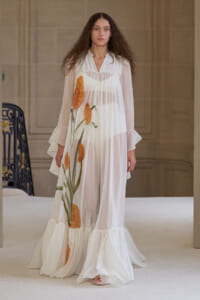 Model on a runway wearing a sheer white, floor-length dress with large orange flowers and a ruffled hem.
