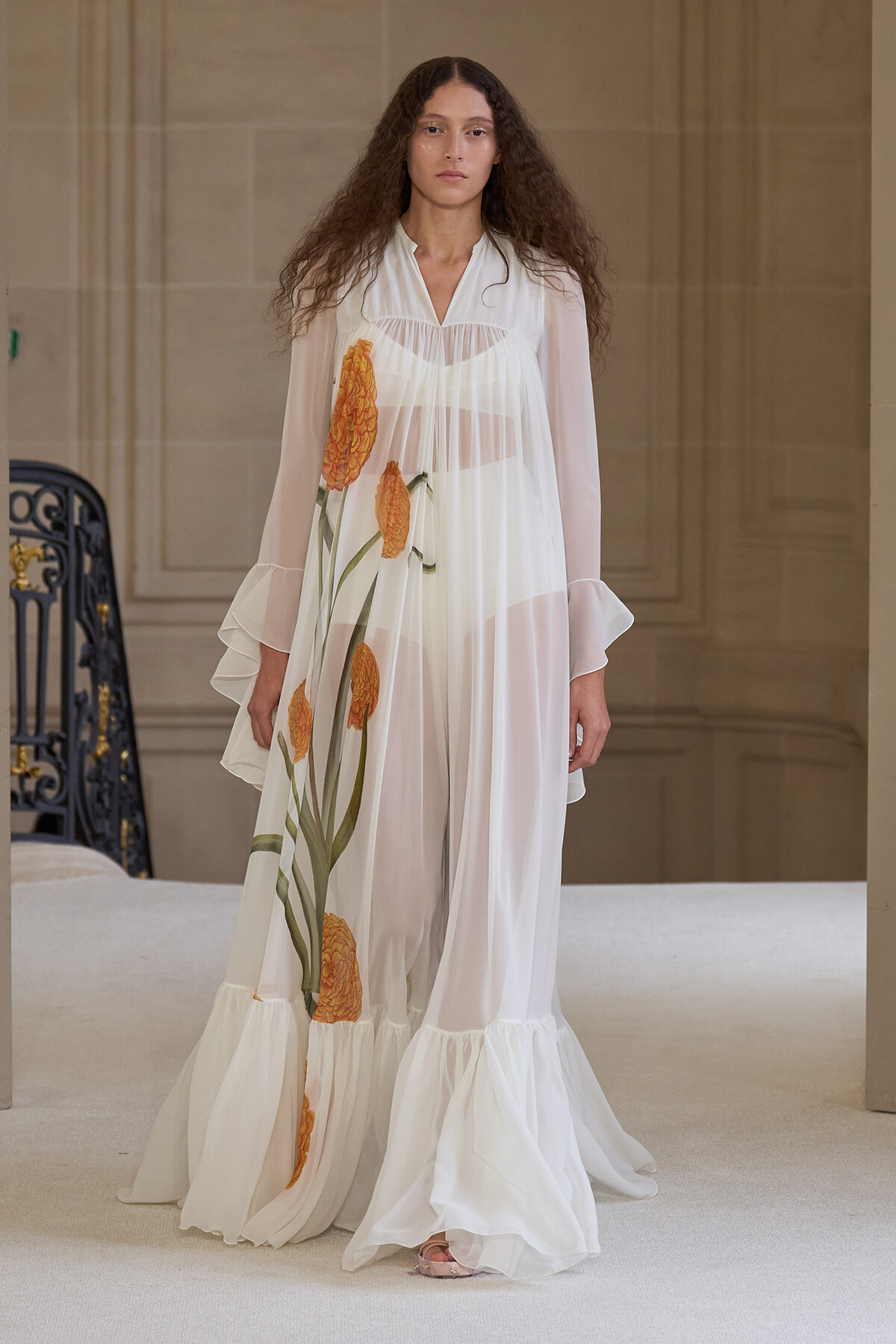 Model on a runway wearing a sheer white, floor-length dress with large orange flowers and a ruffled hem.