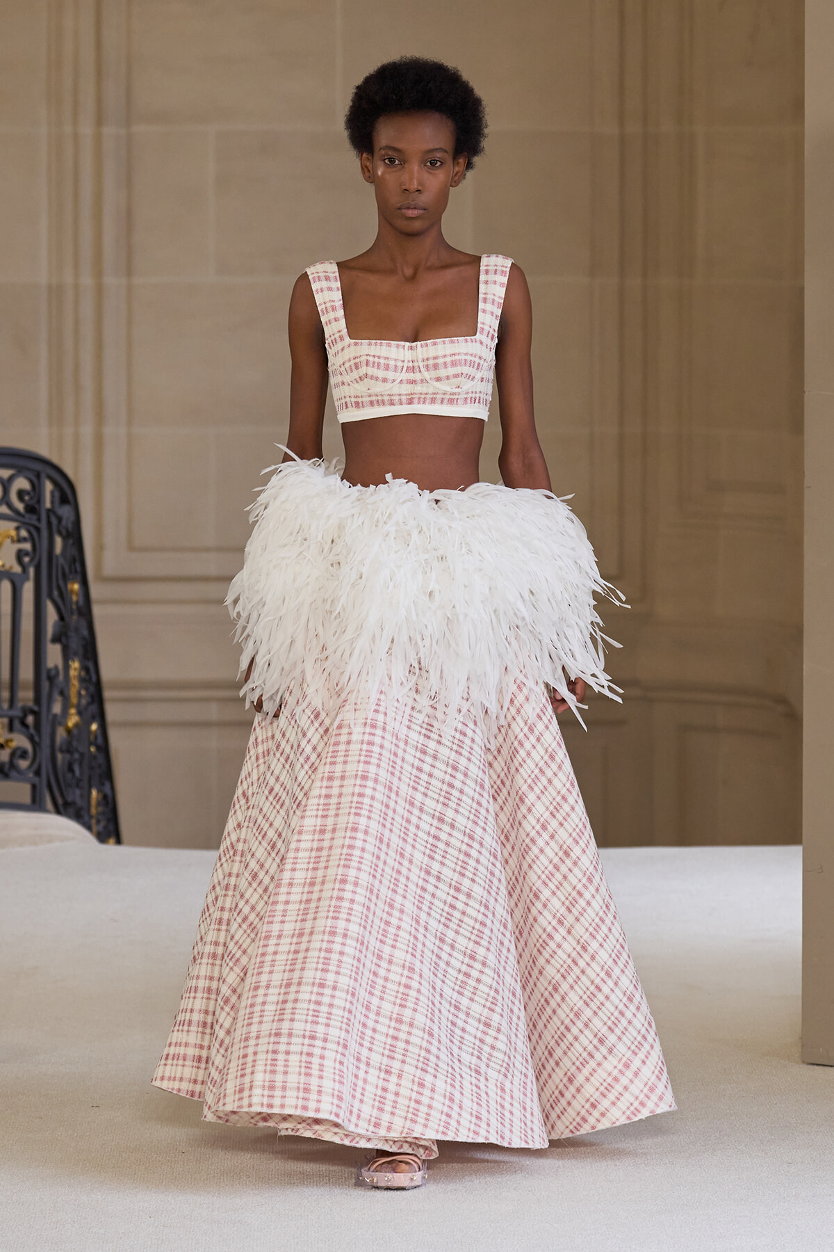 Model walks a fashion runway in a pink plaid two-piece top with a white feather skirt overlay and matching belt.