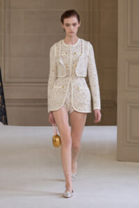 Runway model wearing a cream tweed short suit with white piping and gold buttons, carrying a small gold round handbag.