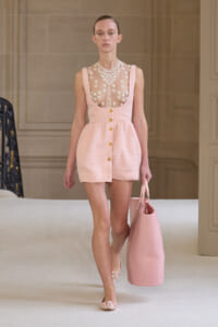 Runway model in a pale pink textured dress with a sheer lace bodice, gold-button front, and a large matching pink tote.