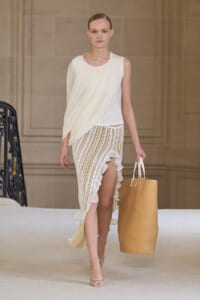 Model walking the runway in a white draped sleeveless top and striped high-low skirt, carrying a large tan tote bag.