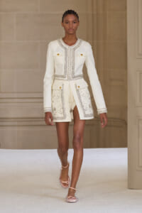 Model walks the runway in an ivory embellished blazer and matching high-waisted shorts, with beaded trim and gold buttons