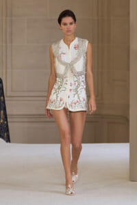 Model on a runway wearing a cream-white, knee-length floral-embroidered mini dress with a heavy beaded vest and open-toe bow-embellished heels.
