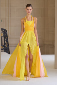 Model wearing a bright yellow gown with pink accents, halter neckline, cinched waist, and long flowing cape-like skirt on a fashion runway