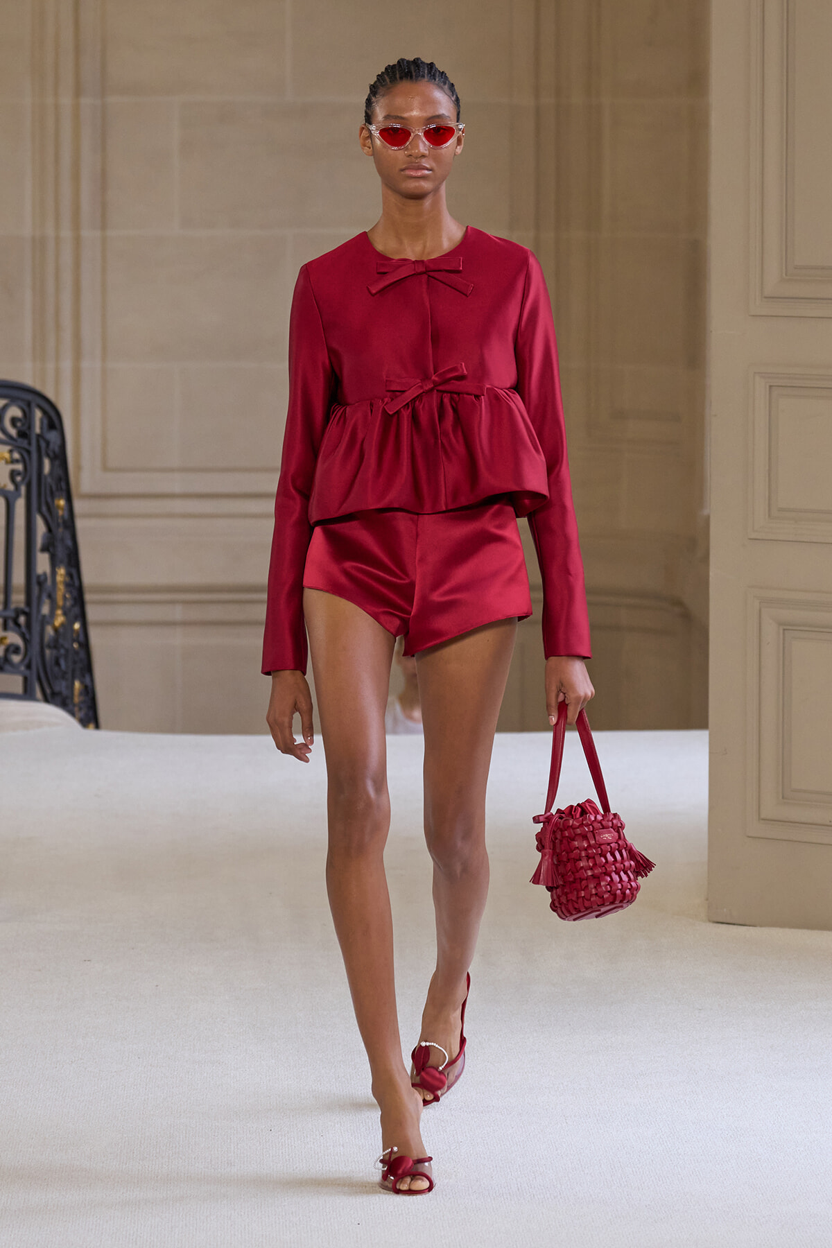Model walking a runway in a burgundy satin romper with a peplum waist, bow neckline, red sunglasses, and matching red handbag and sandals
