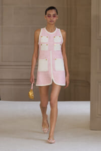 Model walking the runway in a pink sleeveless dress with white pocket panels, paired with pink sunglasses and nude heels, carrying a gold mini bag.