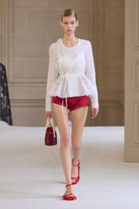 Runway model in a sheer white lace peplum top, red satin shorts, red sandals, and a small red handbag.