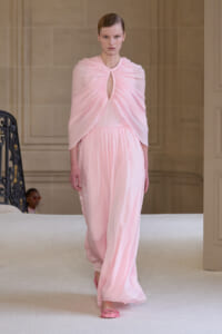 Model wearing a flowing pale pink gown with draped cape walking on a runway.