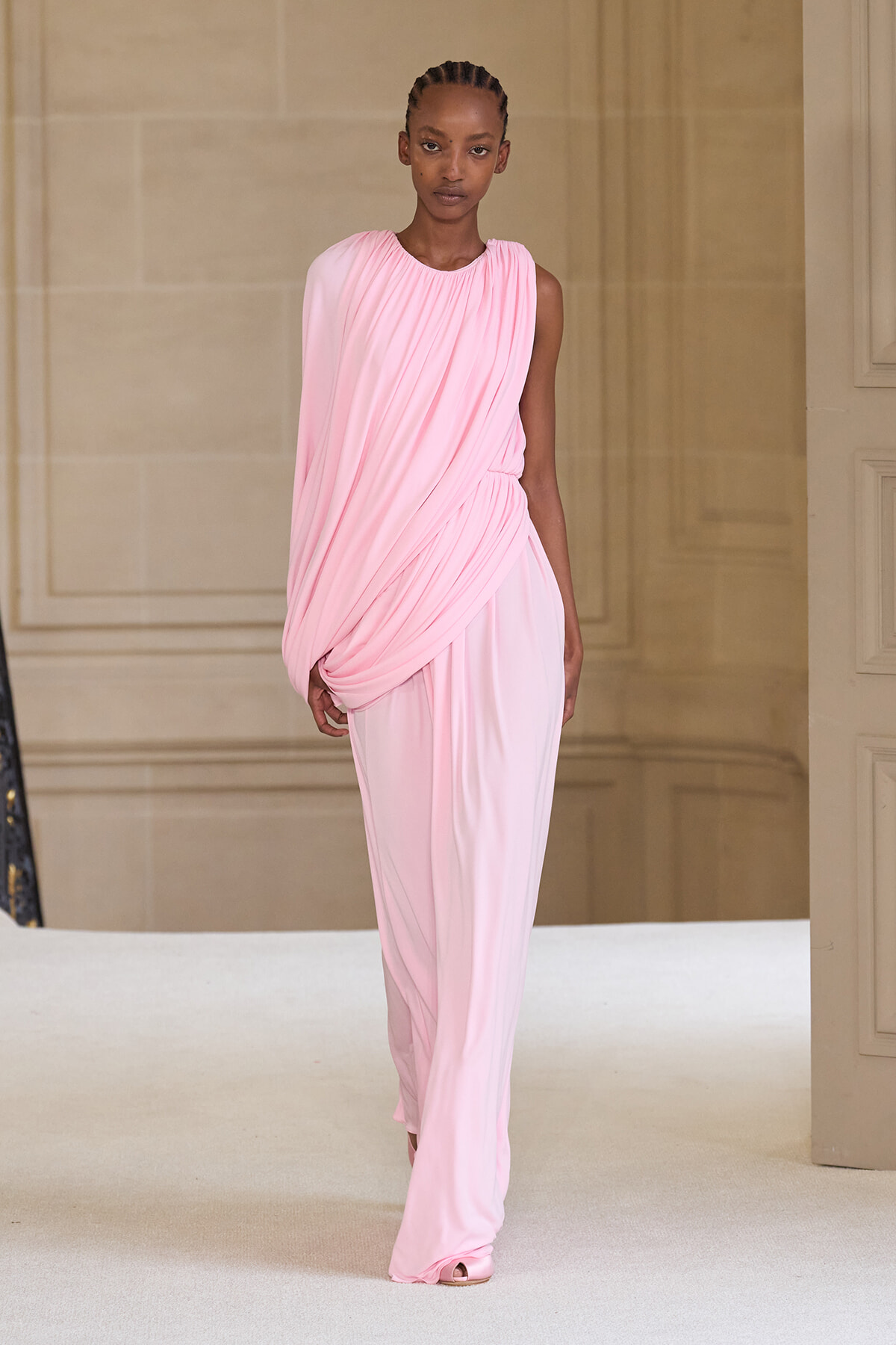 Model walking a runway in a pale pink draped evening gown with one-shoulder design.
