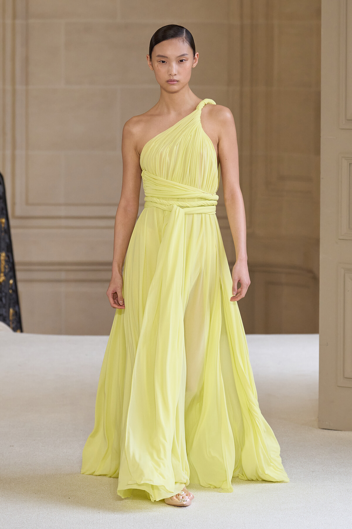Model walking the runway in a pale yellow, one-shoulder chiffon gown with a wrapped bodice and waist sash.