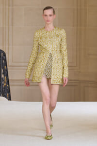 Female model on a runway wearing a gold brocade-patterned long-sleeve coat with a short hem, paired with a shimmering inner dress and olive-green embellished flats.