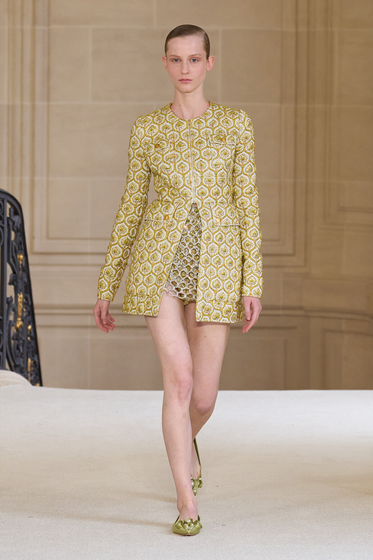 Female model on a runway wearing a gold brocade-patterned long-sleeve coat with a short hem, paired with a shimmering inner dress and olive-green embellished flats.