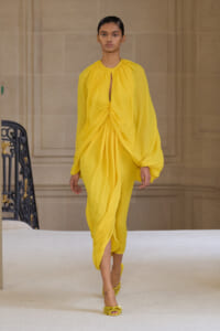 Model walks a runway in a bright yellow draped midi dress with a gathered center knot and long bishop sleeves, paired with yellow heeled sandals.