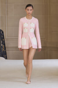 Model on a runway wearing a pink tweed coat with white pocket panels and gold buttons, mid-stride.