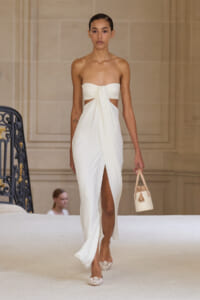 Model walking a runway in a white strapless dress with a twisted knot at the bust and a high leg slit, holding a small beige handbag.