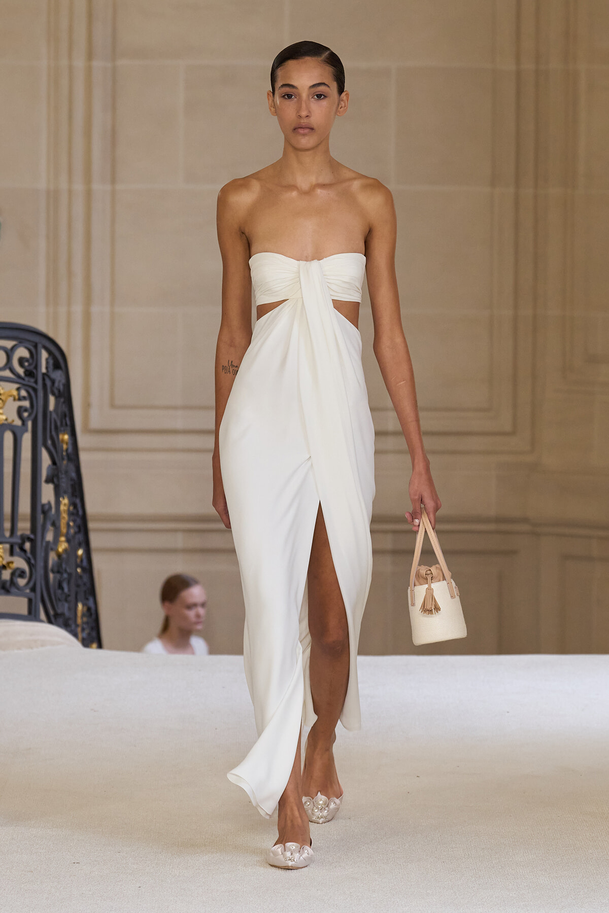 Model walking a runway in a white strapless dress with a twisted knot at the bust and a high leg slit, holding a small beige handbag.