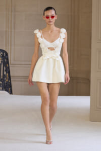 Model walking a runway in a white structured mini dress with flower appliques along the straps and a bow at the waist, wearing red sunglasses.