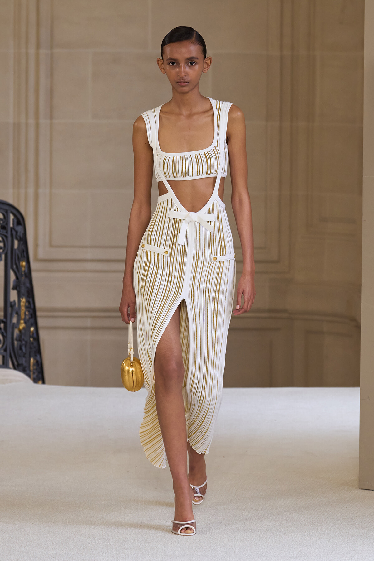 Model walking a runway in a white and gold striped cutout dress with a bow belt and side pockets, carrying a small gold handbag.