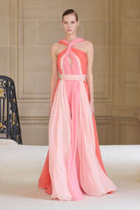 Model on a runway wearing a floor-length pink gradient gown with a twisted halter neckline, soft waist sash, and flowing chiffon panels.