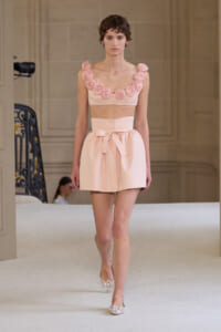 Model walks the runway in a pale pink two-piece outfit: a cropped top with pink fabric roses along the neckline and a high-waisted skirt with a bow at the waist, paired with pink flats.