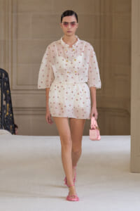 Model walking runway in a white, embroidered mini dress with pink floral accents and pink accessories.