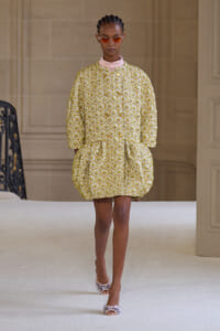 Model walking a runway in a cropped gold-and-white patterned bubble dress with a pink high-neck underneath and orange sunglasses.