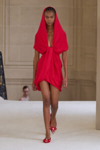 Runway model wearing a bright red draped hooded mini dress and matching red embellished heels, walking forward on a catwalk.